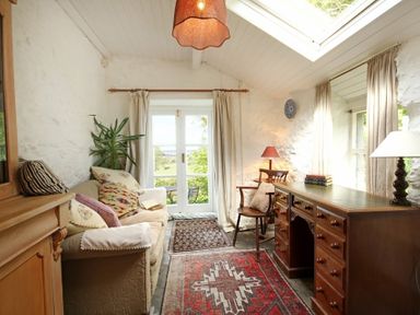 A sitting room with a sofa multiple cushions a wooden desk and chair with lamps and rugs at Buck Cottage in Red Wharf Bay