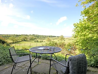 A patio with a round metal table and two chairs overlooking green fields and trees at Buck Cottage in Red Wharf Bay