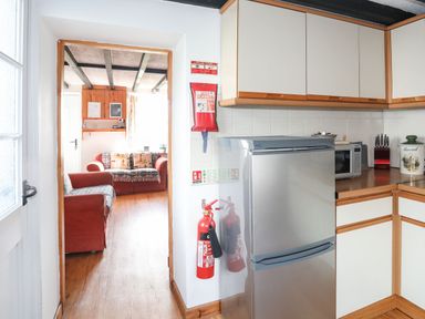 A kitchen with a silver fridge microwave knives and bread container visible next to a living room with red sofas and wooden beams at Cae Person in Llangoed