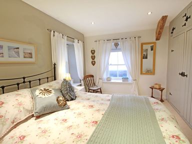 A bedroom with a floral bedspread, pillows, a chair, a small side table, a window with white curtains, a mirror, and built-in wardrobes at Cae'r Dyni (Caer Dyni) in Criccieth
