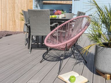 An outdoor patio with grey wicker dining chairs around a table and a pink circular lounge chair next to a plant and an open book with a green apple on it at Cambrian House in Criccieth