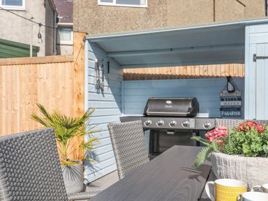 An outdoor dining area with a table, wicker chairs, a potted plant, a barbecue grill, and cooking utensils at Cambrian House in Criccieth