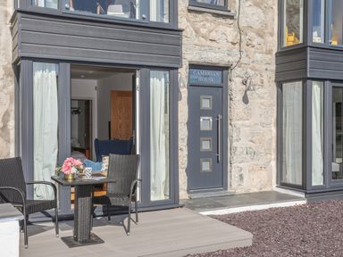 A patio area with two black chairs and a table with flowers outside a stone building with a door labeled Cambrian House in Criccieth