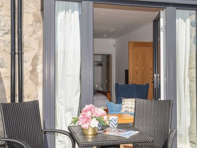 An outdoor seating area with two woven chairs and a table with flowers and mugs in front of open glass doors showing a room with a blue armchair and bed at Cambrian House in Criccieth