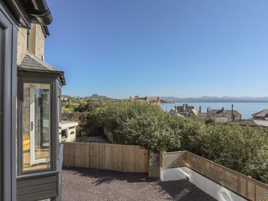 A view of a fenced backyard and bushes with houses and water in the distance at Cambrian House in Criccieth