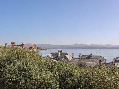 Houses with rooftops behind bushes with a body of water and mountains in the background at Cambrian House in Criccieth