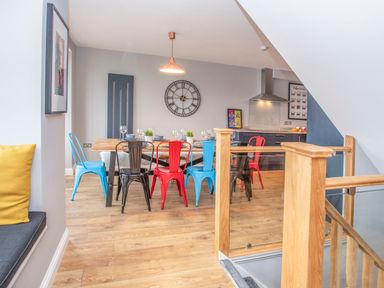 A dining area with a wooden table and multicolored metal chairs next to a kitchen and wooden staircase at Cambrian House in Criccieth