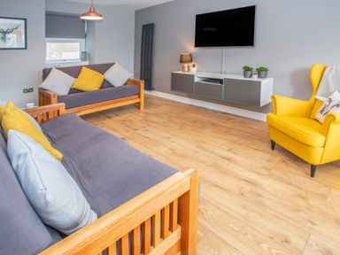 A living room with two wooden sofas with cushions a yellow armchair a wall-mounted television and a floating cabinet with plants and a lamp at Cambrian House in Criccieth