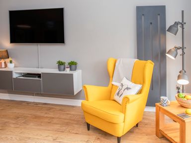 A yellow armchair with a pillow and throw in a living room with a wall-mounted TV and plants on a cabinet at Cambrian House in Criccieth