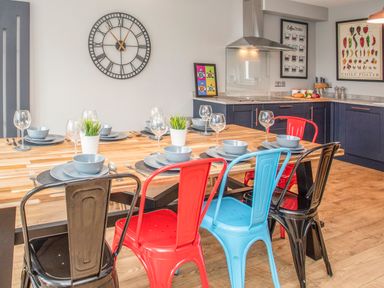 A dining area with a wooden table set with plates bowls wine glasses and plants metal chairs in red blue and black in a kitchen at Cambrian House in Criccieth