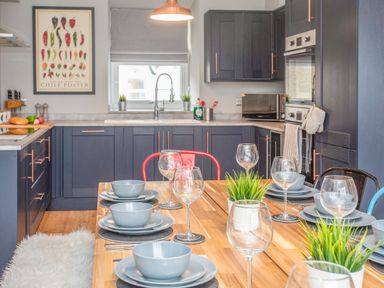A kitchen with blue cabinets a wooden dining table set with plates bowls wine glasses and plants at Cambrian House in Criccieth