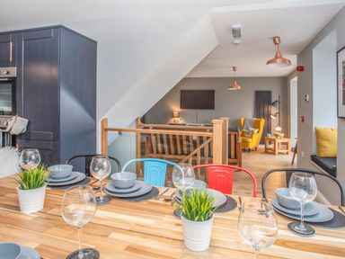 A dining table set with glassware and plates in an open plan kitchen and living area with colorful chairs and a yellow armchair at Cambrian House in Criccieth