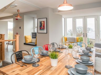 A dining area with a wooden table set with grey dishes and glassware and colorful chairs at Cambrian House in Criccieth