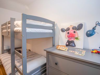 A bedroom with gray bunk beds a gray chest of drawers a cow painting domino game and a blue desk lamp at Cambrian House in Criccieth