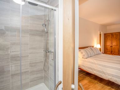 A bedroom with a bed striped bedding and a wooden wardrobe seen through a partially open door next to a tiled shower enclosure at Cambrian House in Criccieth