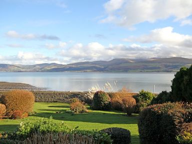 A garden with bushes and grass overlooking a body of water and hills in the background at Camlan in Beaumaris