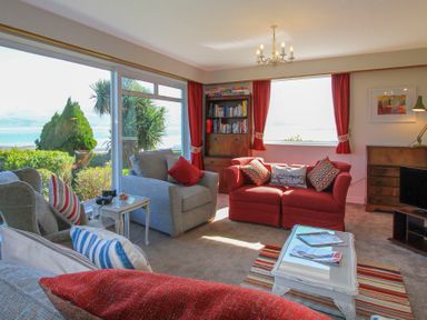 A living room with sofas and armchairs by large windows with a view of the sea and garden at Camlan in Beaumaris