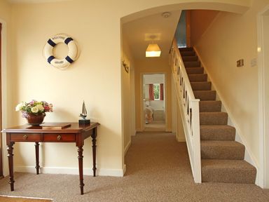 A hallway with a wooden table holding flowers and a decorative boat under a life ring on the wall and stairs to the right at Camlan in Beaumaris