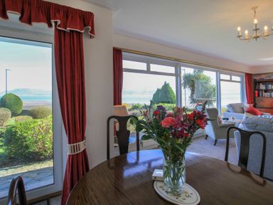 A dining table with a vase of flowers near large windows and red curtains in a living room at Camlan in Beaumaris