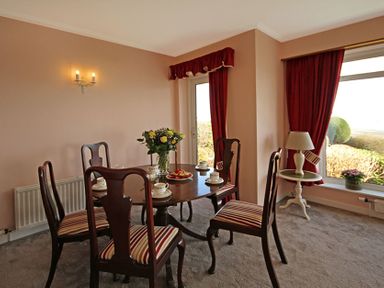 A dining room with a wooden table and striped chairs with red curtains on windows at Camlan in Beaumaris