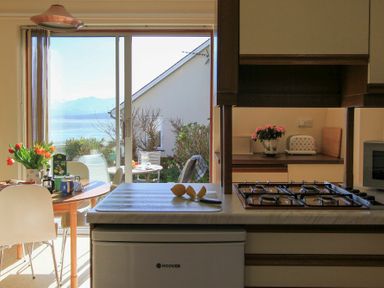 A kitchen with a gas stove and cut lemons on the counter next to a dining table with flowers and mugs near sliding glass doors showing an outdoor seating area and water view at Camlan in Beaumaris