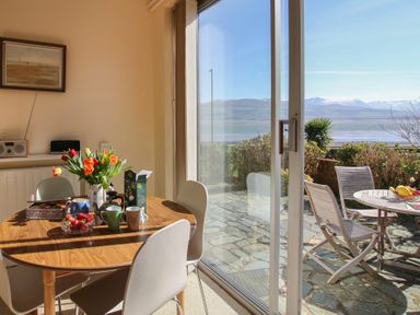 A dining area with a wooden table set with cups and flowers next to a glass door opening to a patio with outdoor chairs and a table overlooking water and mountains at Camlan in Beaumaris