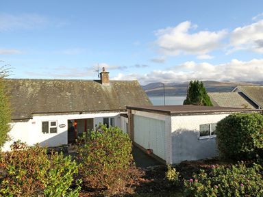 A white house with a chimney and an attached garage surrounded by bushes with hills and a body of water in the background at Camlan in Beaumaris