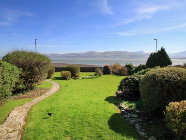 A garden with a stone path and trimmed bushes near water with mountains in the background at Camlan in Beaumaris
