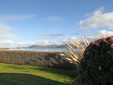 A garden with flowers and reeds by a stone wall overlooking water and mountains at Camlan in Beaumaris