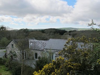 A stone house with a slate roof surrounded by trees and yellow flowering bushes in a rural area