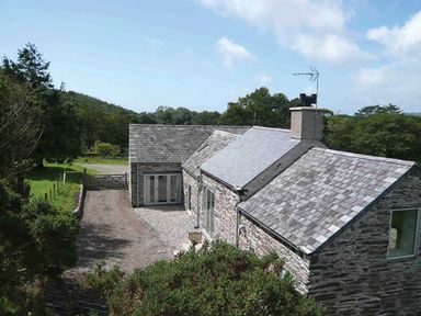 A stone house with a slate roof surrounded by trees and a gravel driveway at Cefn Isaf in Porthmadog