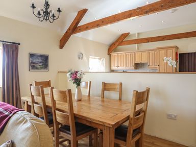 A dining area with wooden table and chairs and flower vase open kitchen with wooden cabinets and exposed ceiling beams at Tan Twr - Chellow Cottage in Dwyran