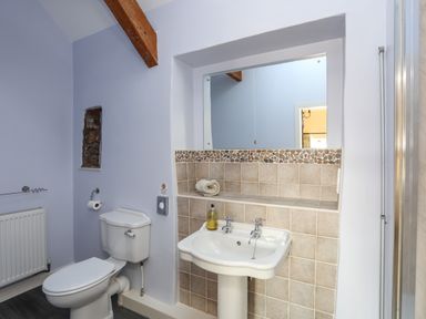 A bathroom with a toilet, pedestal sink, tiled wall, mirror, and exposed wooden beam at Tan Twr - Chellow Cottage in Dwyran