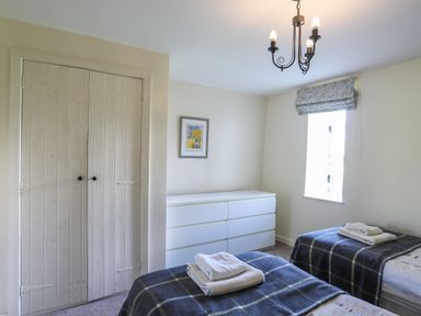 A bedroom with two single beds with folded towels a white dresser a window with a patterned blind and a closed wooden closet at Tan Twr - Chellow Cottage in Dwyran