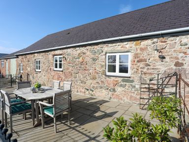An outdoor patio area with a wooden dining table and chairs next to a stone building at Tan Twr - Chellow Cottage in Dwyran