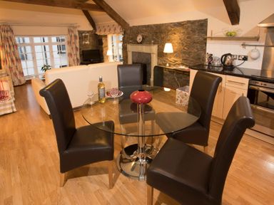 A dining area with a round glass table and four chairs next to a kitchen and living room at The Coach House Beaumaris