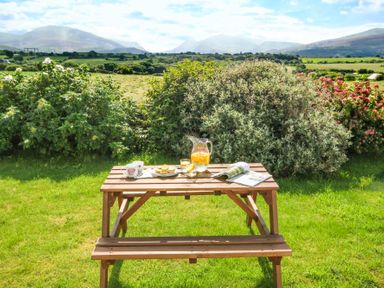 A wooden picnic table with mugs plates a jug of juice and a magazine in a garden with bushes and mountains in the background at Coed Bolyn Lodge in Bethel