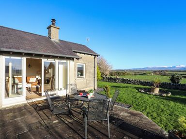 An outdoor patio with a metal table and four chairs next to a house with glass doors opening to a living room and green fields with mountains in the background at Cosy Barn in Llangaffo