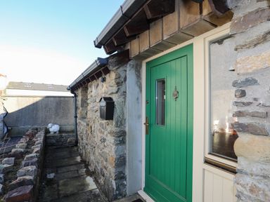 A stone exterior wall with a green door and a black mailbox beside a small stone path at Cosy Barn in Llangaffo