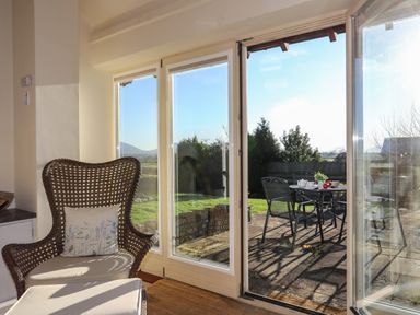 A wicker chair with a cushion next to glass doors opening to a patio with a table and metal chairs at Cosy Barn in Llangaffo