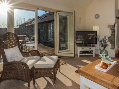 A sitting area with a wicker chair, ottoman, a television on a stand, and a wooden coffee table in a room with glass doors leading outside at Cosy Barn in Llangaffo