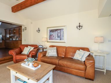 A living room with a brown leather sofa and armchair a wooden coffee table with decorations and a kitchen area in the background at Cosy Barn in Llangaffo