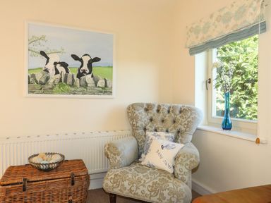 A patterned armchair with cushions beside a window with a vase of flowers and a wicker basket with decorative balls in a room at Cosy Barn in Llangaffo