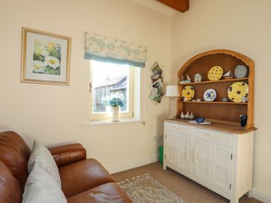 A sofa with cushions next to a window and a cabinet with decorative plates and a lamp at Cosy Barn in Llangaffo