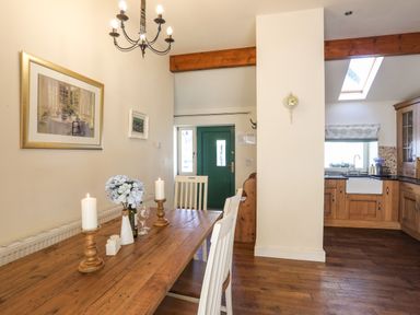 A dining area with wooden table white chairs candles and flowers next to a kitchen with wooden cabinets and a green door at Cosy Barn in Llangaffo