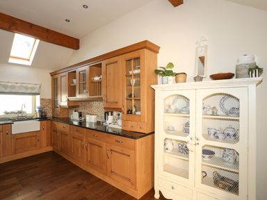 A kitchen with wooden cabinets and a white display cabinet showcasing dishes at Cosy Barn in Llangaffo