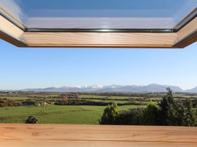 A landscape view of green fields and snow capped mountains seen through an open wooden window at Cosy Barn in Llangaffo