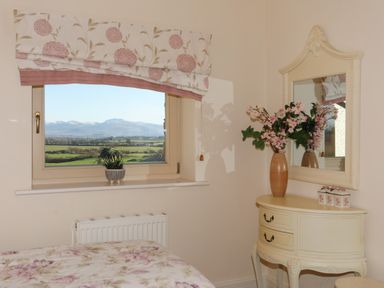 A bedroom with a window showing a landscape and a small table with a mirror and flowers at Cosy Barn in Llangaffo