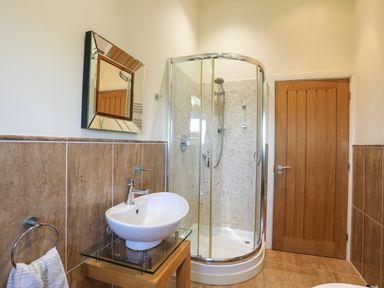 A bathroom with a glass shower enclosure a wooden door a white sink on a glass and wood stand and a mirror above the sink at Cosy Barn in Llangaffo