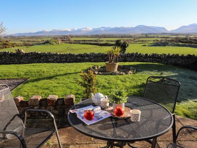 A patio with metal table and chairs and a teapot with cups overlooking a green lawn and stone wall with mountains in the background at Cosy Barn in Llangaffo